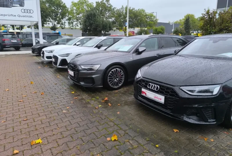 Pet Audi vozila parkirano u nizu na placu auto salona, crni model u prvom planu, mokar asfalt sa jesenjim lišćem nakon kiše