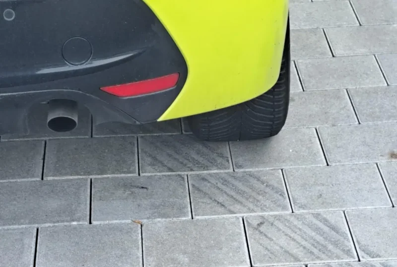 Detalj stražnjeg branika žutog automobila s ispušnom cijevi i crvenim reflektorom parkiranog na sivom popločanom parkingu peugeot 208 hdi